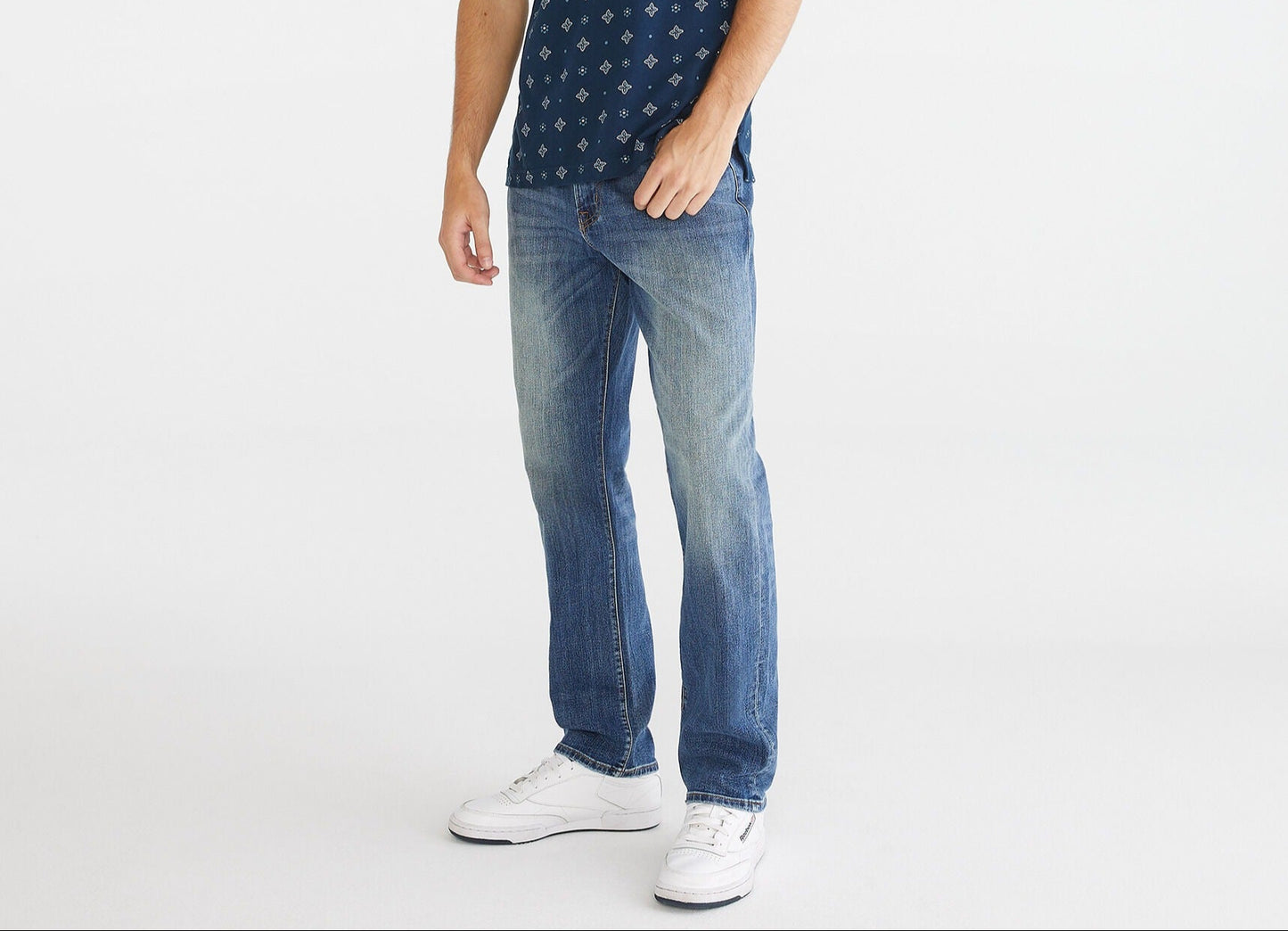 Man wearing a navy patterned polo shirt and blue jeans on a white background