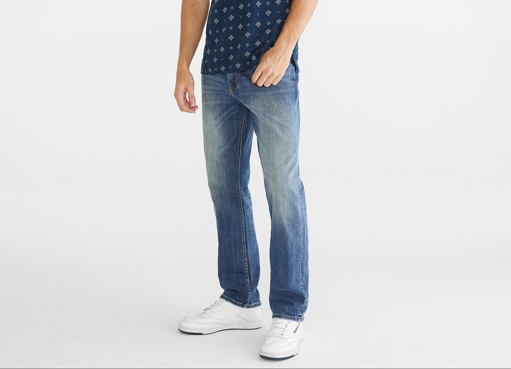 Man wearing a navy patterned polo shirt and blue jeans on a white background