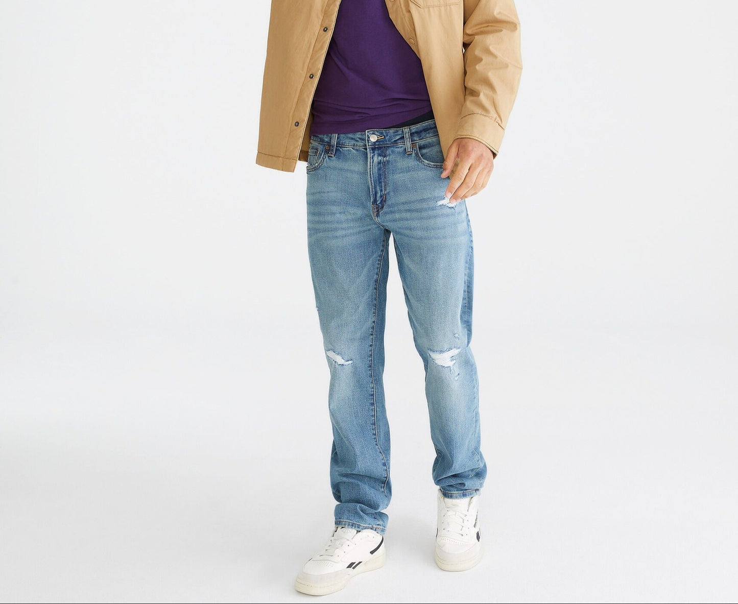 Man wearing a tan jacket, purple shirt, and blue jeans on a white background
